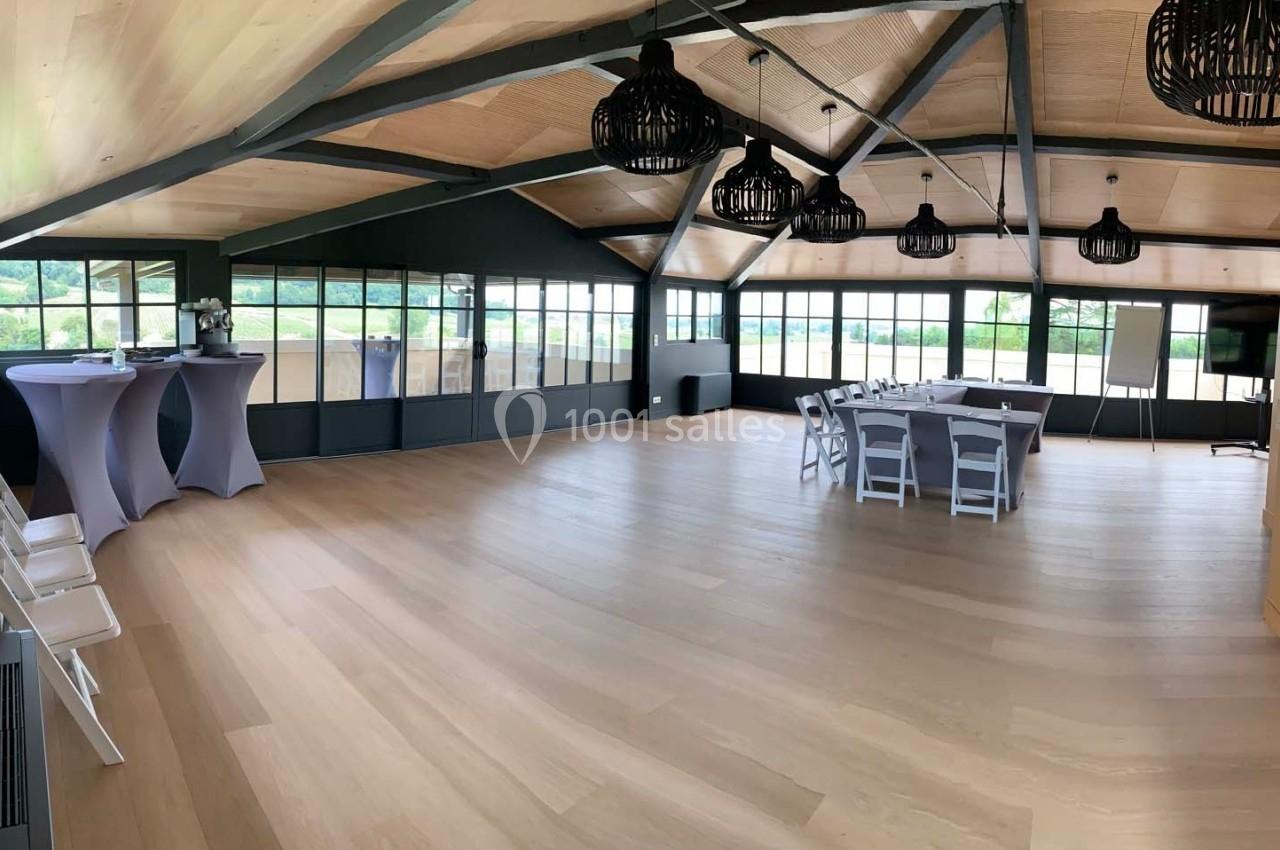 Salle lumineuse avec parquet clair, grandes baies vitrées, tables et chaises blanches, et plafond en bois avec poutres…
