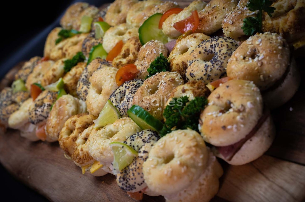 Plateau de mini-bagels garnis de légumes, fromage et charcuterie, disposés sur une planche en bois.
