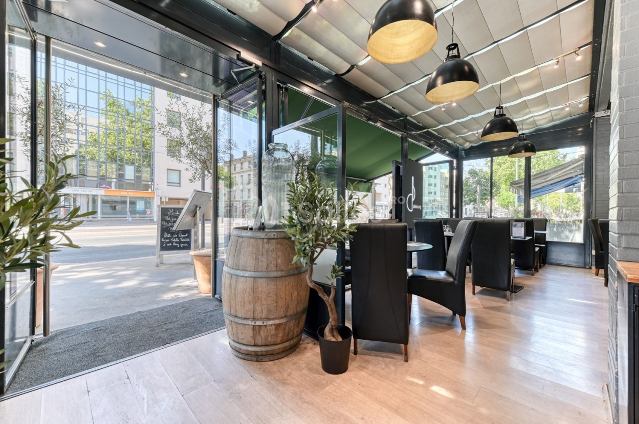Salle de restaurant lumineuse avec baies vitrées, mobilier moderne, tonneau en bois et plantes décoratives.