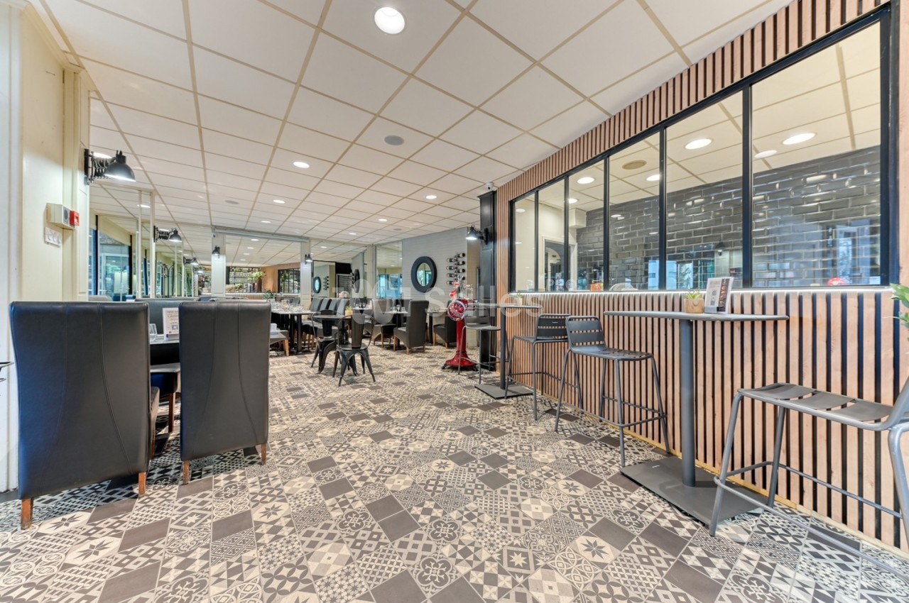 Salle de restaurant moderne avec chaises noires, tables en bois, sol à motifs et cloison vitrée.