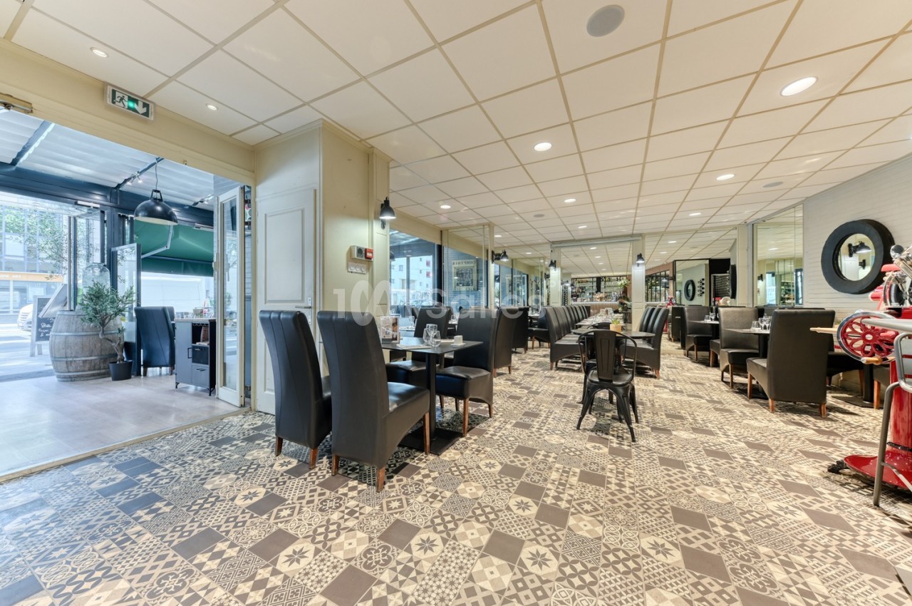 Salle de restaurant lumineuse avec tables dressées, chaises noires et sol en carreaux de ciment décoratifs.