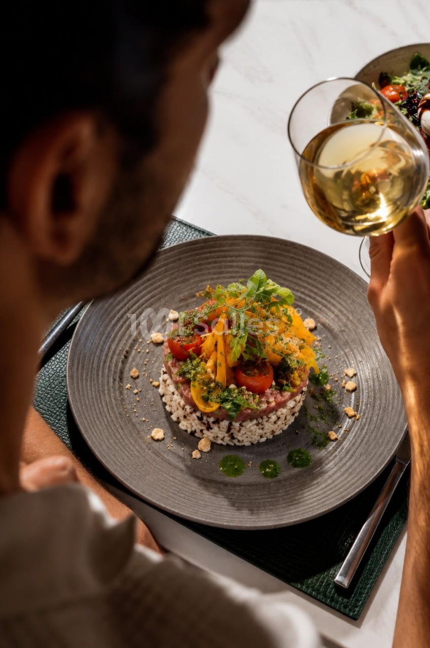 Convive tenant une assiette grise avec risotto, légumes et sauce, verre de vin blanc à la main.