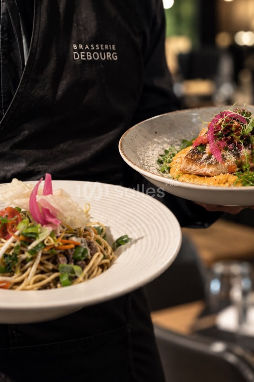Serveur en vêtement noir présente deux assiettes - noodles aux légumes et poisson grillé aux herbes potagères.
