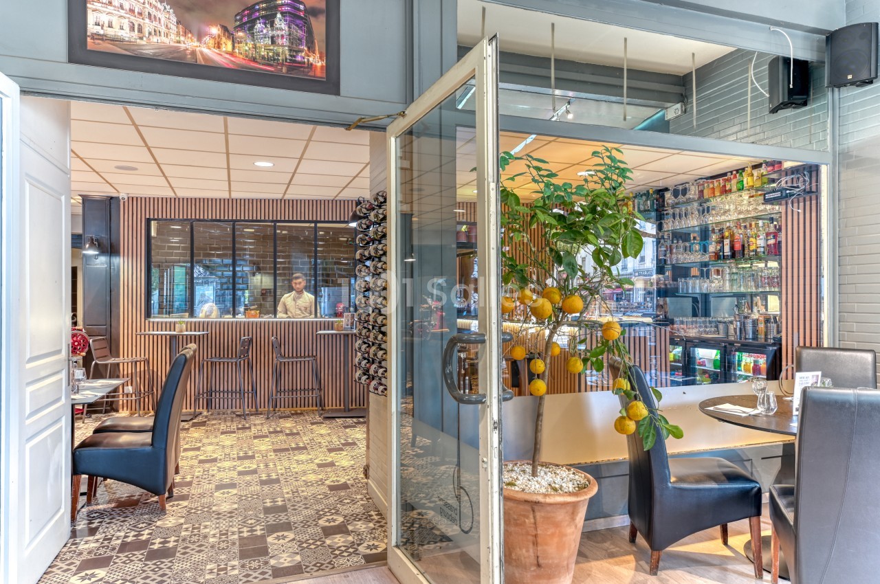 Intérieur de brasserie avec comptoir en bois, tables dressées, chaises bleues et décoration végétale aux agrumes.