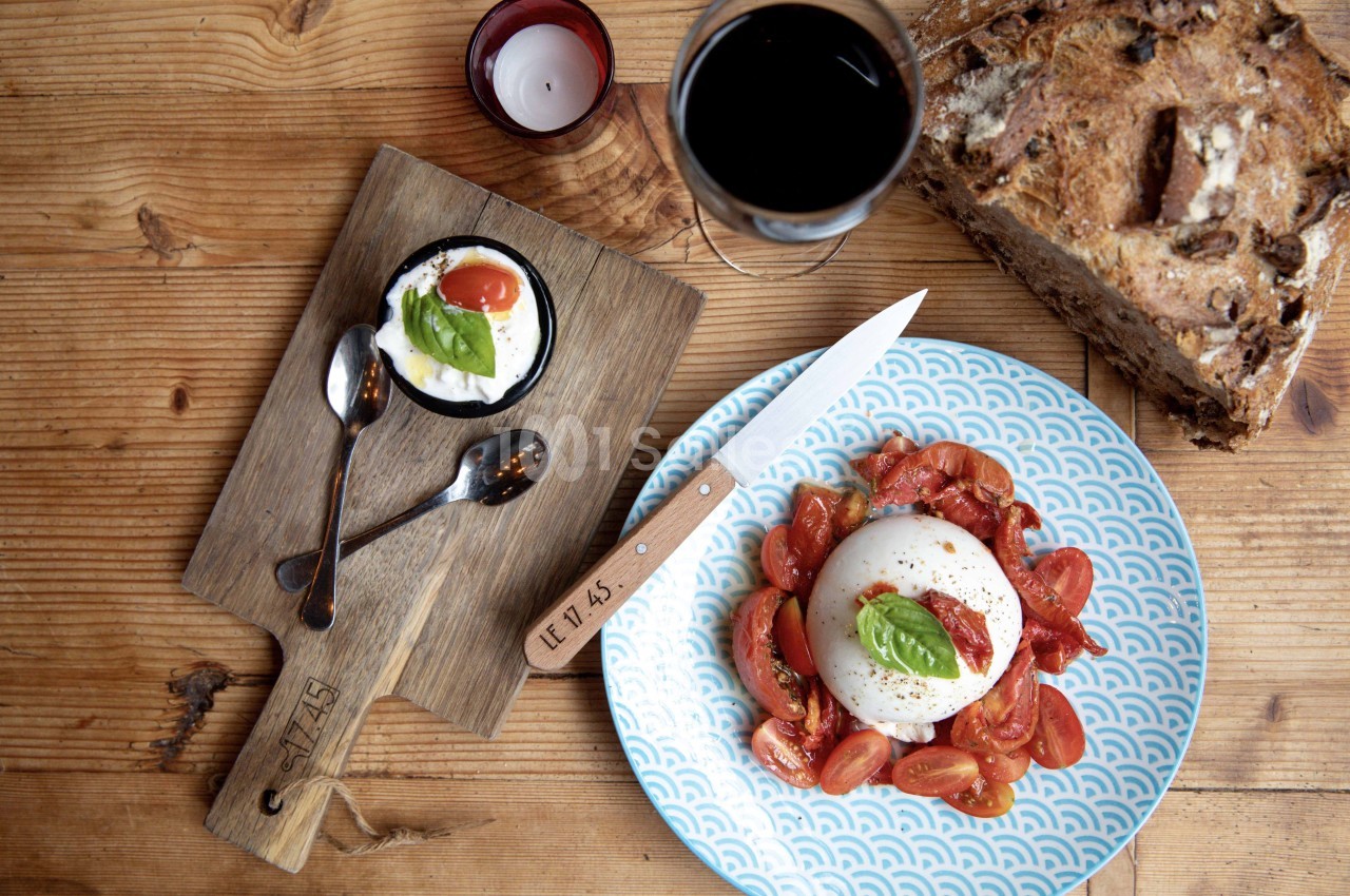 Assiette de burrata avec tomates cerises et basilic, accompagnée de pain, verre de vin et dessert sur une table en bois.