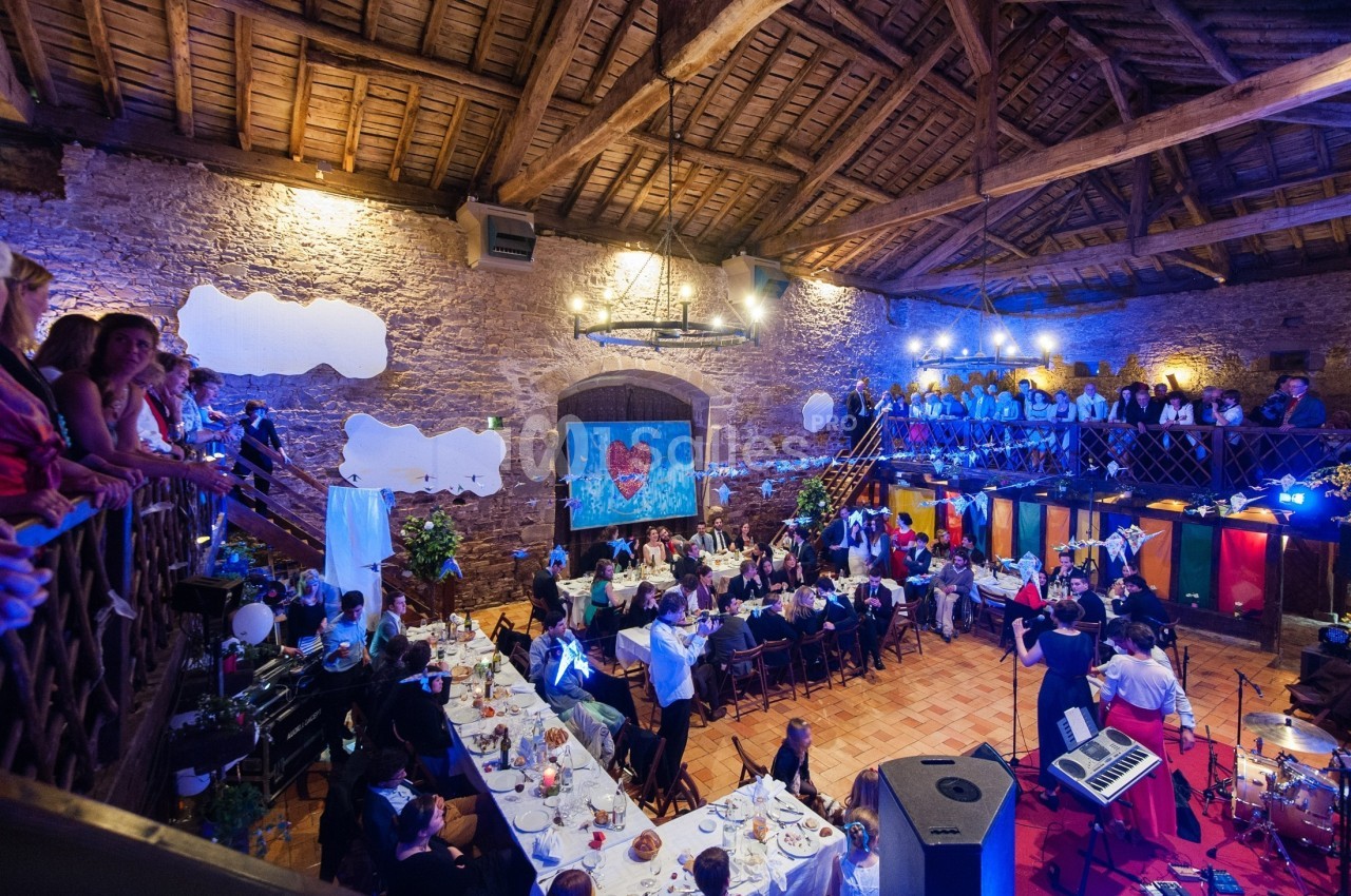 Salle en pierre avec plafond en poutres, tables dressées pour un repas, invités assis et musiciens sur scène.