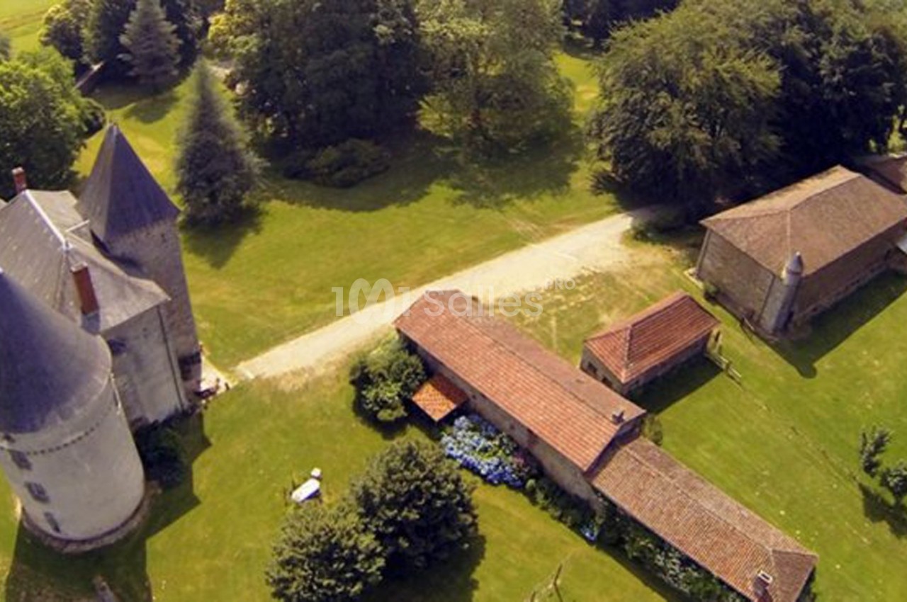 Vue aérienne d'un château avec tours rondes, entouré de bâtiments annexes et de verdure en milieu rural.