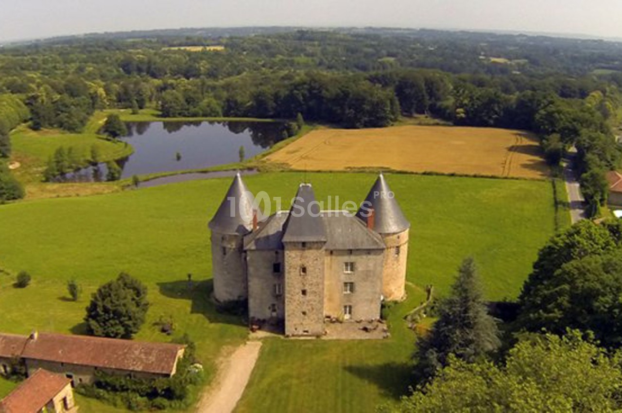 Vue aérienne d'un château entouré de champs, d'un étang et de bâtiments annexes dans un paysage rural verdoyant.