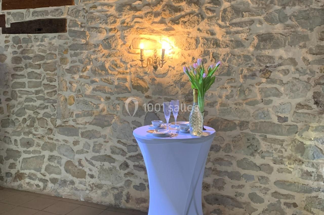 Table haute avec nappe blanche, décorée de tulipes dans un vase et de coupes, devant un mur en pierre éclairé par une…