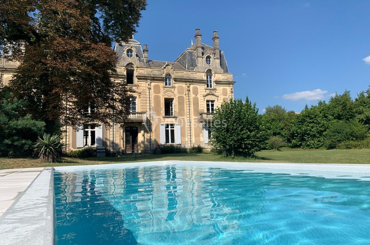 Façade d'un château en pierre entouré de verdure, avec une piscine au premier plan sous un ciel bleu.