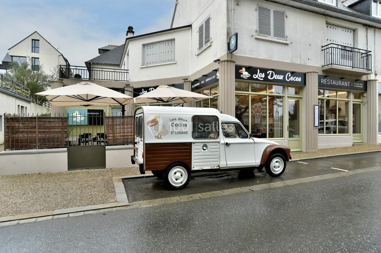 Une voiture vintage blanche et marron stationnée devant un restaurant avec terrasse et parasols.