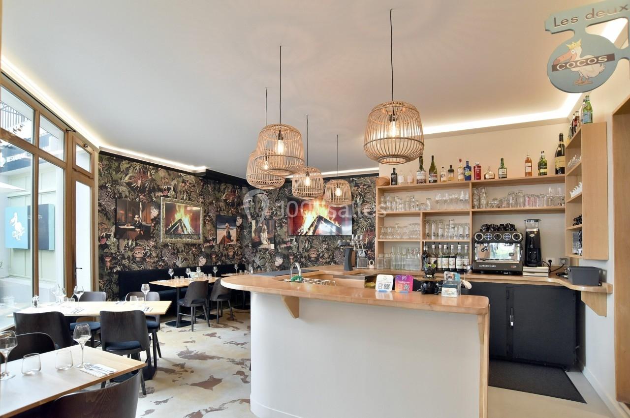 Intérieur d'un restaurant avec un comptoir en bois, des luminaires suspendus et des tables dressées près d'une baie vitrée.