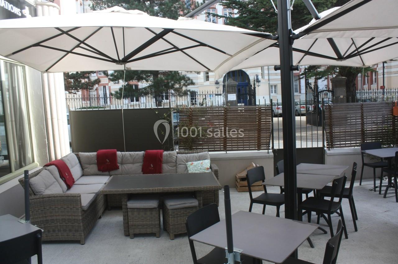 Terrasse aménagée avec canapés en rotin, tables et chaises sous de grands parasols, entourée de panneaux et végétation.
