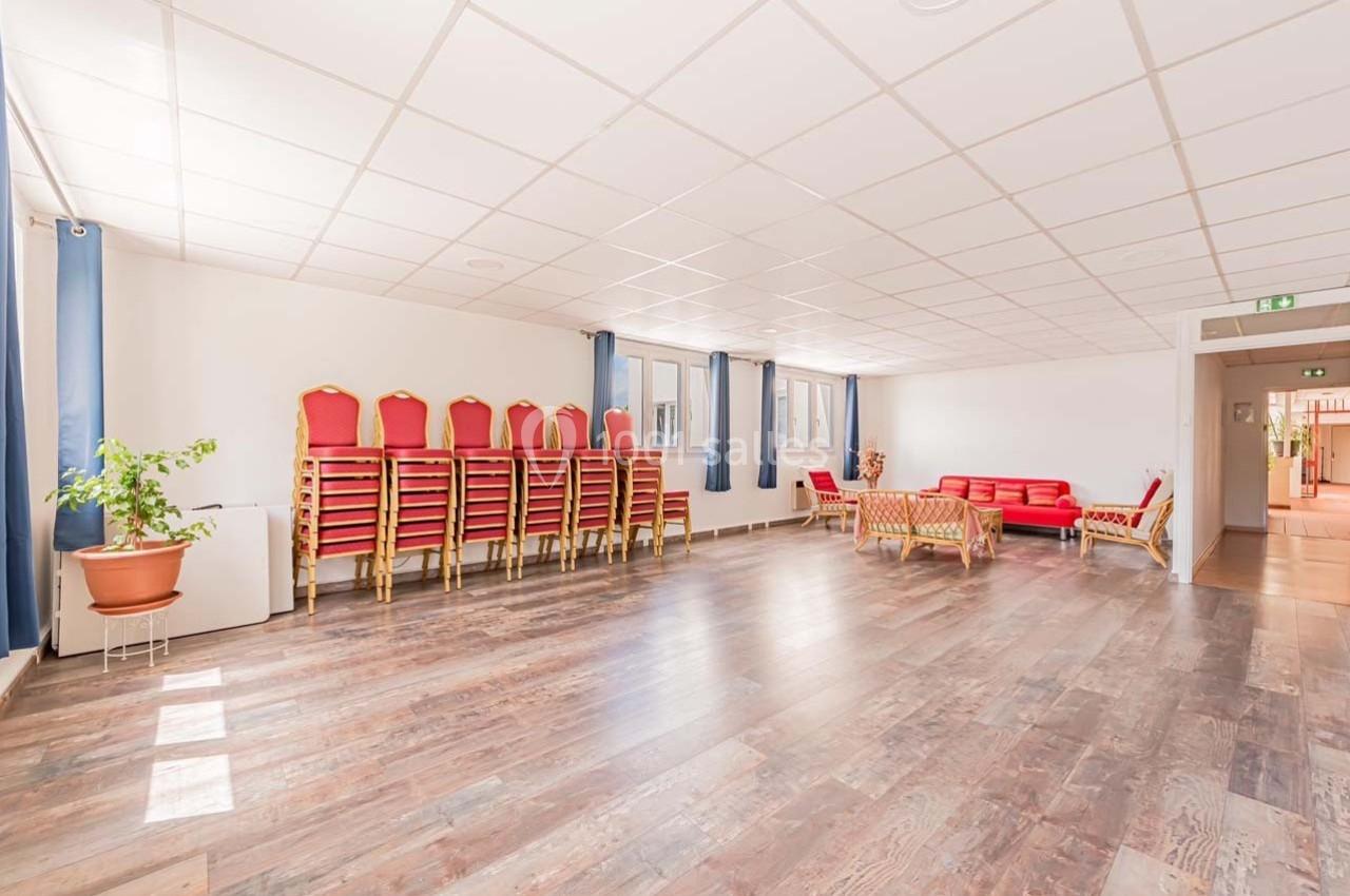 Salle lumineuse avec parquet, chaises empilées contre un mur, canapés rouges, plantes et grandes fenêtres.