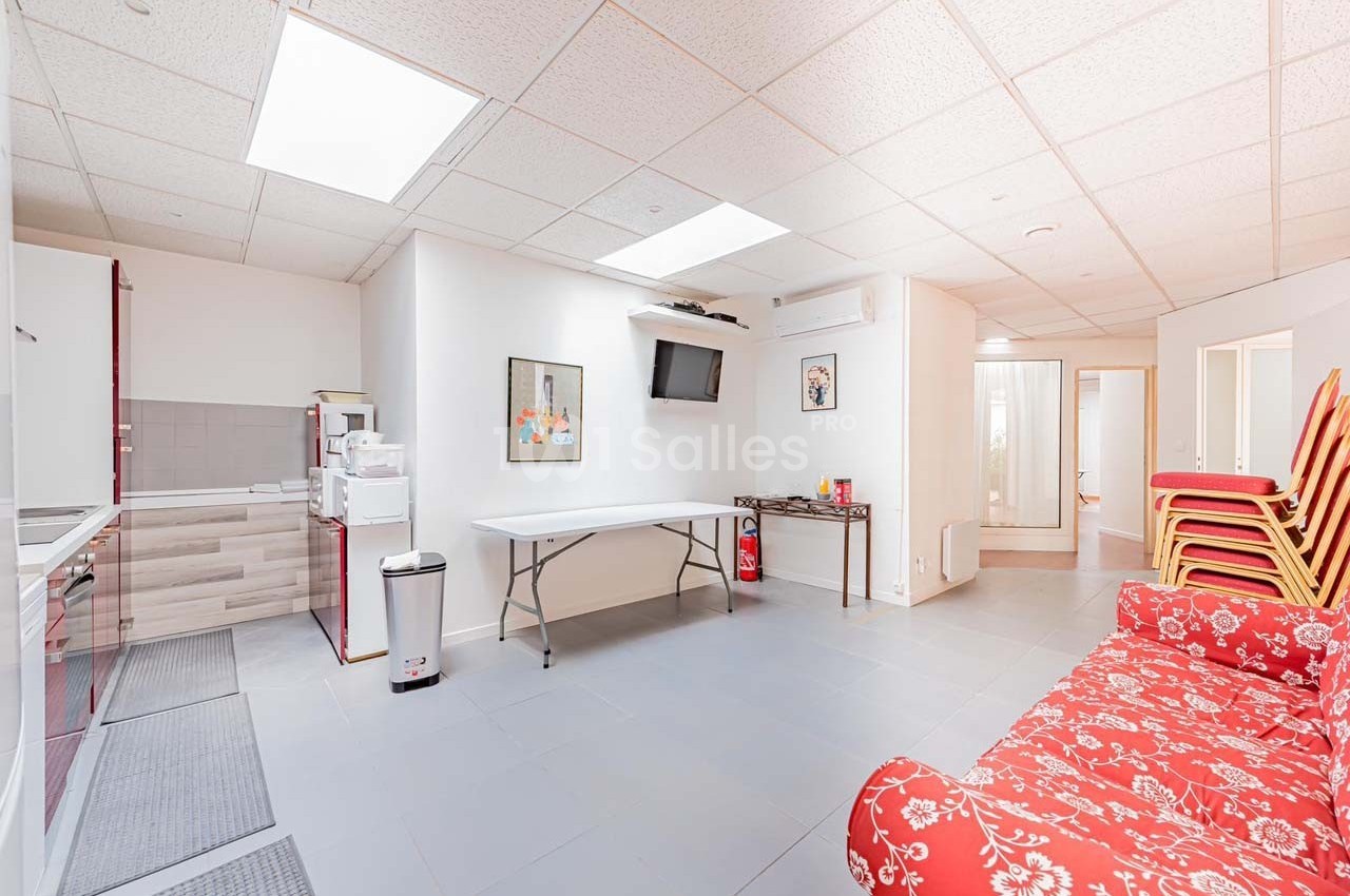 Salle lumineuse avec canapé rouge à motifs, table pliante, chaises empilées, kitchenette et écran mural.