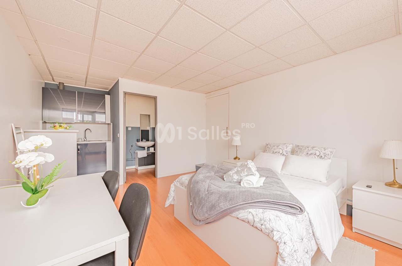 Chambre lumineuse avec lit double, bureau, kitchenette et salle de bain attenante dans un espace moderne et épuré.