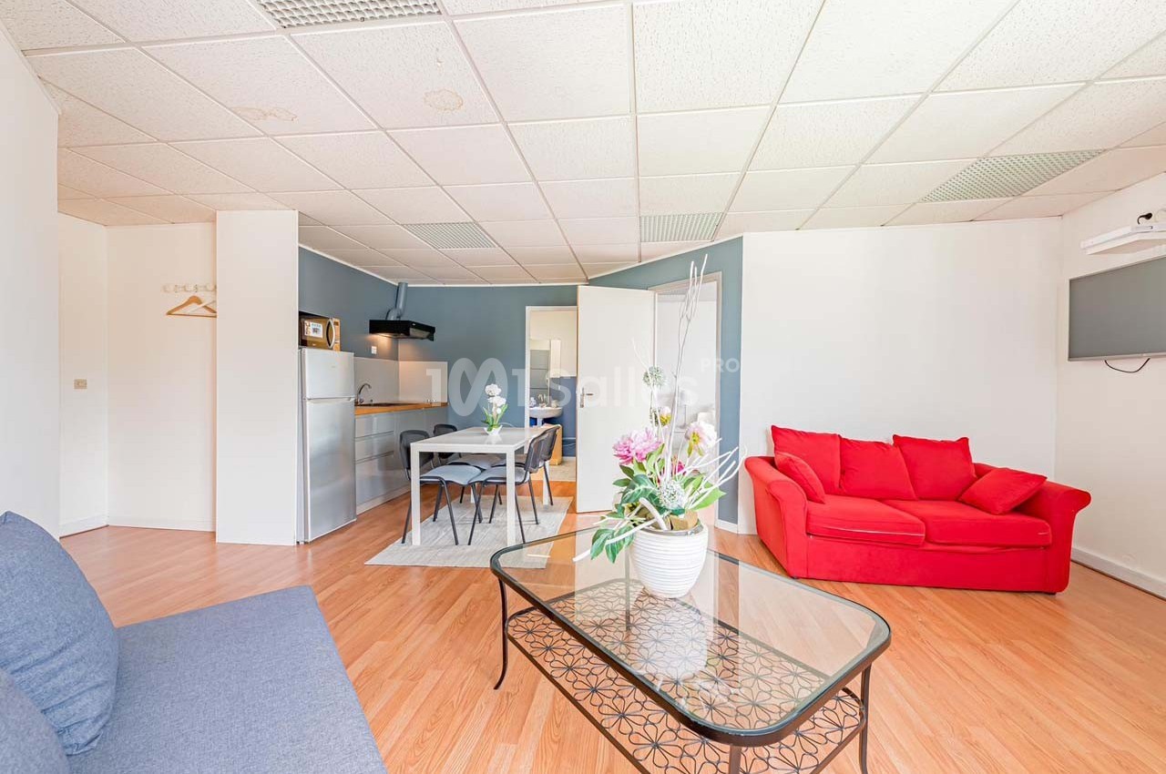 Salon lumineux avec canapé rouge, table basse en verre, coin repas et cuisine ouverte en arrière-plan.