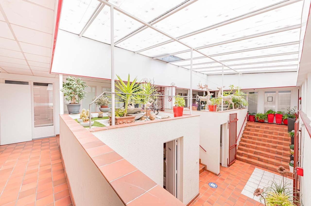 Cour intérieure lumineuse avec toit en verre, plantes en pots et escaliers en carrelage rouge.