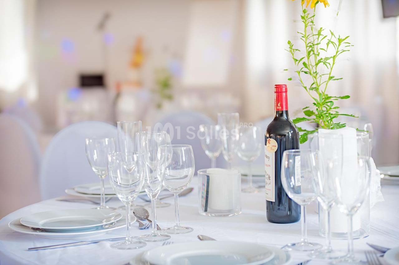 Table élégamment dressée avec nappes blanches, verres, assiettes et une bouteille de vin rouge au centre.