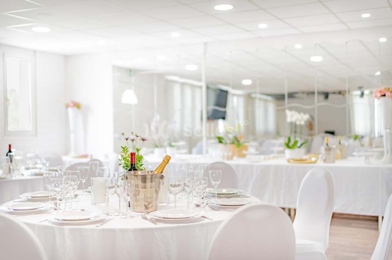 Salle de réception lumineuse avec tables rondes dressées, décorations élégantes et seau à champagne au premier plan.