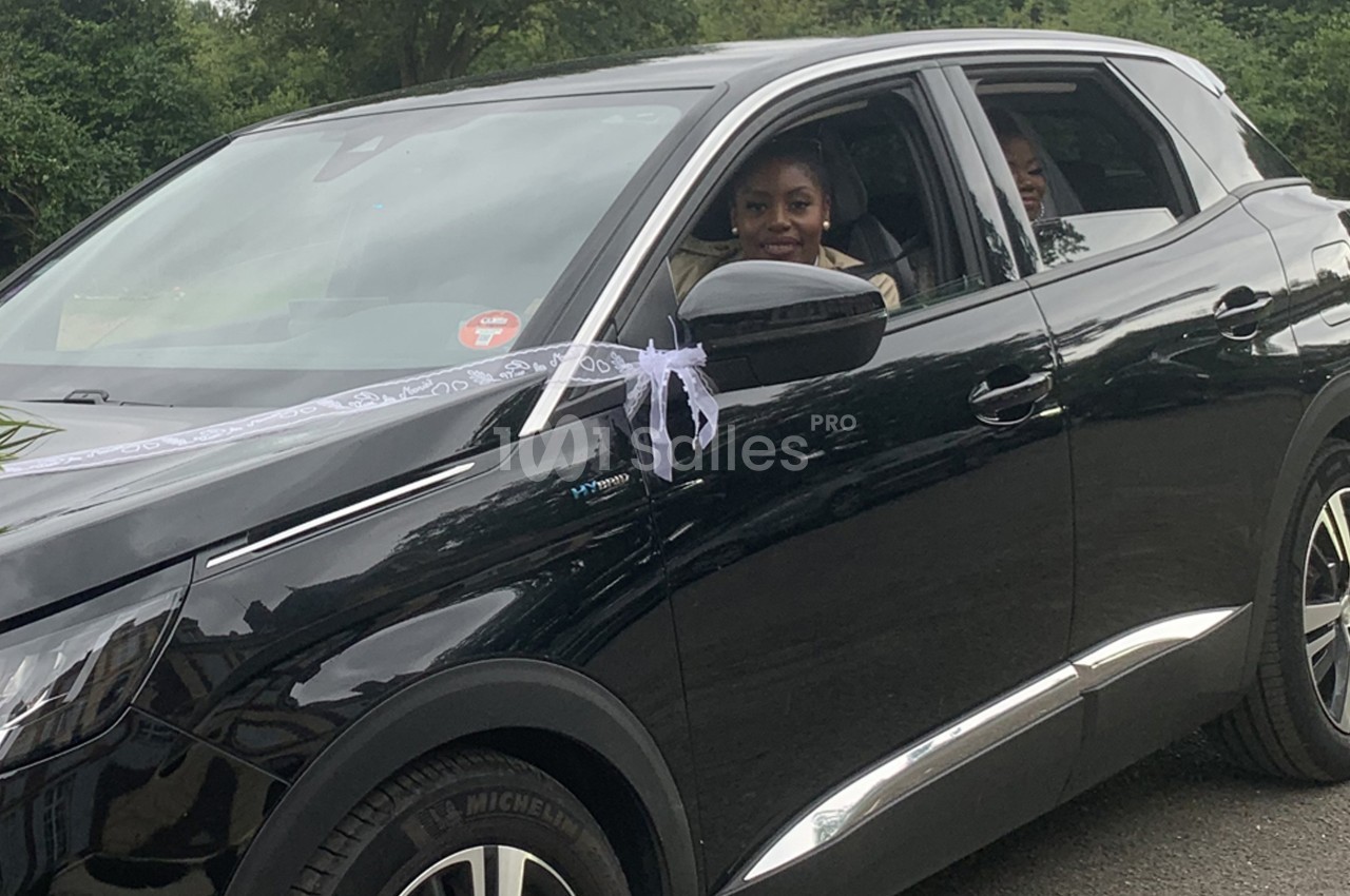 Une femme souriante dans une voiture noire décorée d'un ruban blanc, stationnée sur une route bordée de verdure.