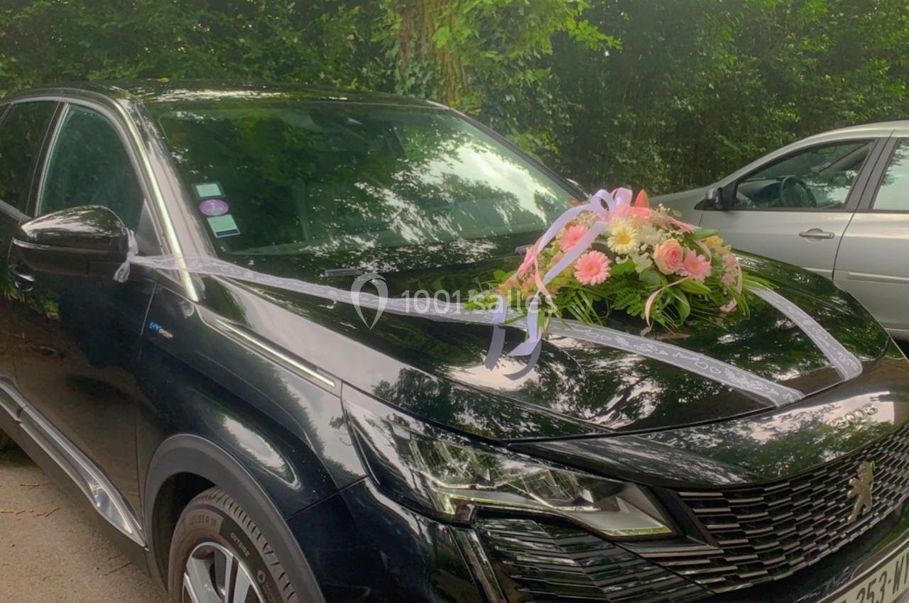 Voiture noire décorée de rubans blancs et d'une composition florale rose sur le capot, stationnée en extérieur.