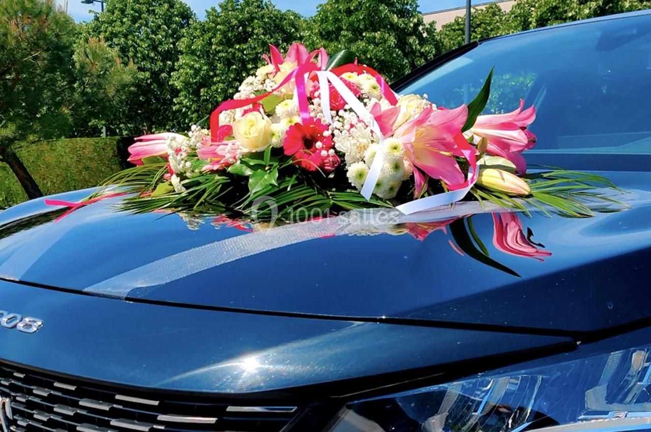 Bouquet floral coloré avec rubans blancs posé sur le capot d'une voiture noire, entourée de verdure.