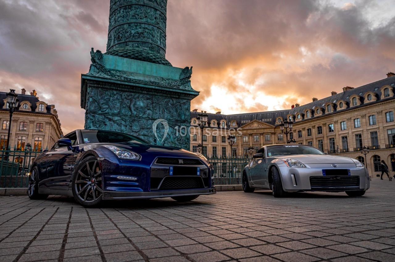 Deux voitures de sport garées sur une place pavée devant une colonne monumentale et des bâtiments historiques au coucher du…
