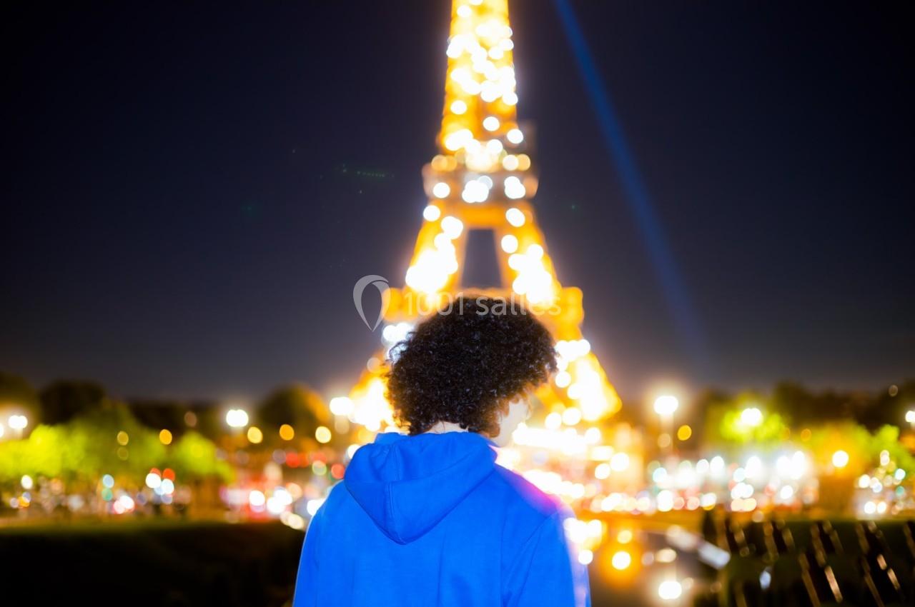 Une personne de dos en sweat bleu regarde la Tour Eiffel illuminée de nuit, entourée de lumières urbaines.