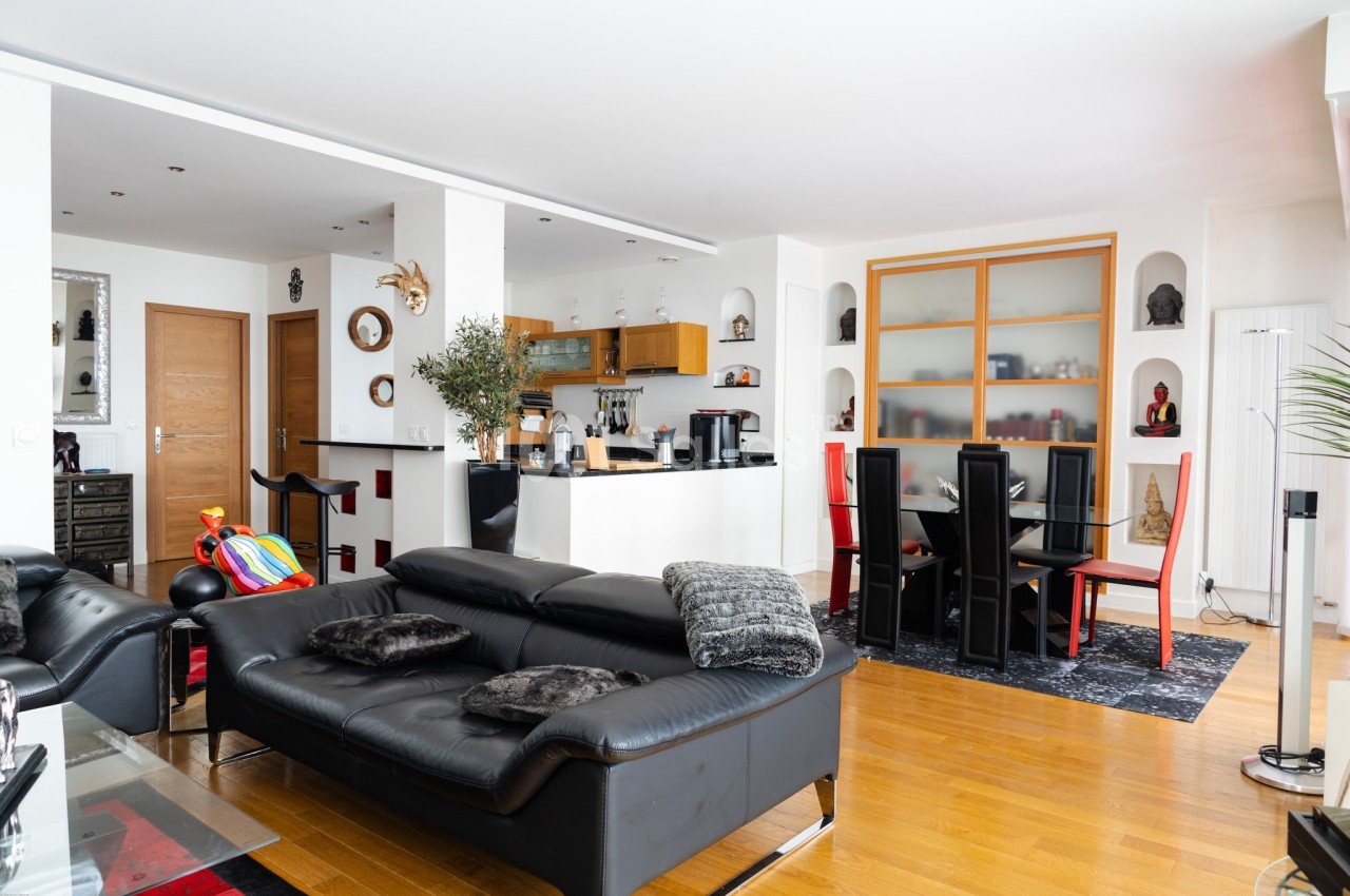 Salon moderne avec canapé en cuir noir, table à manger, cuisine ouverte et parquet clair.