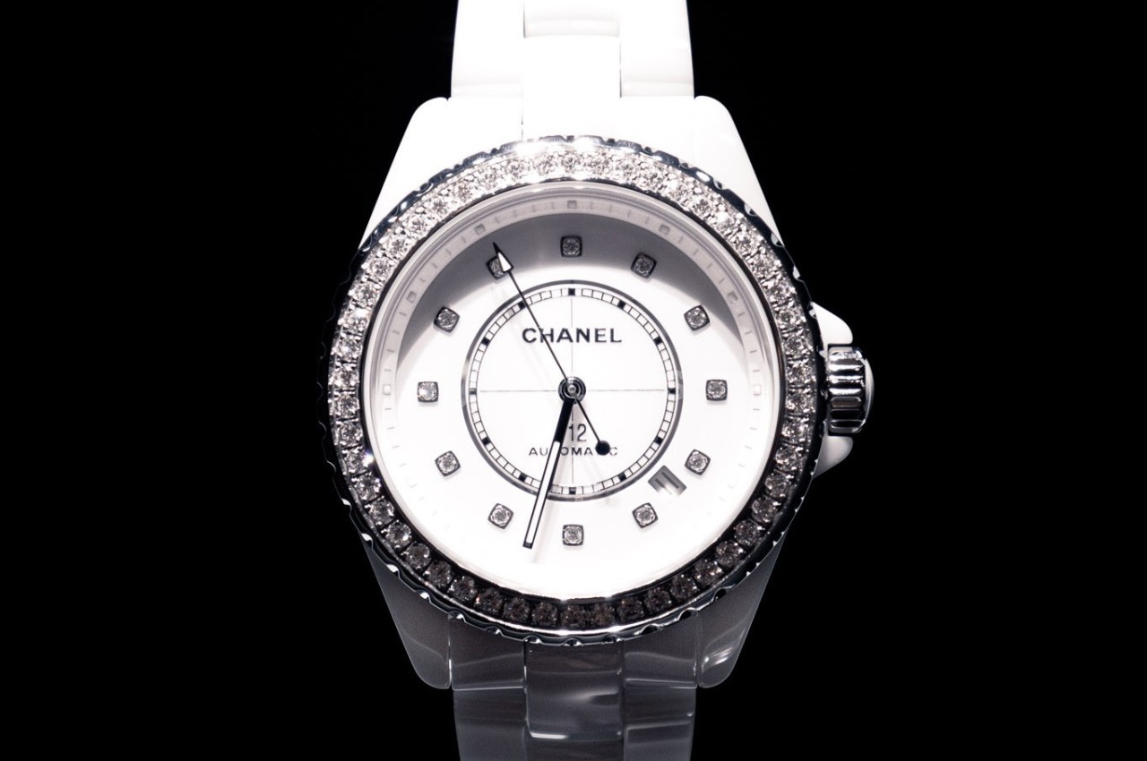 Montre blanche avec cadran rond, index en diamants et lunette sertie de pierres brillantes sur fond noir.
