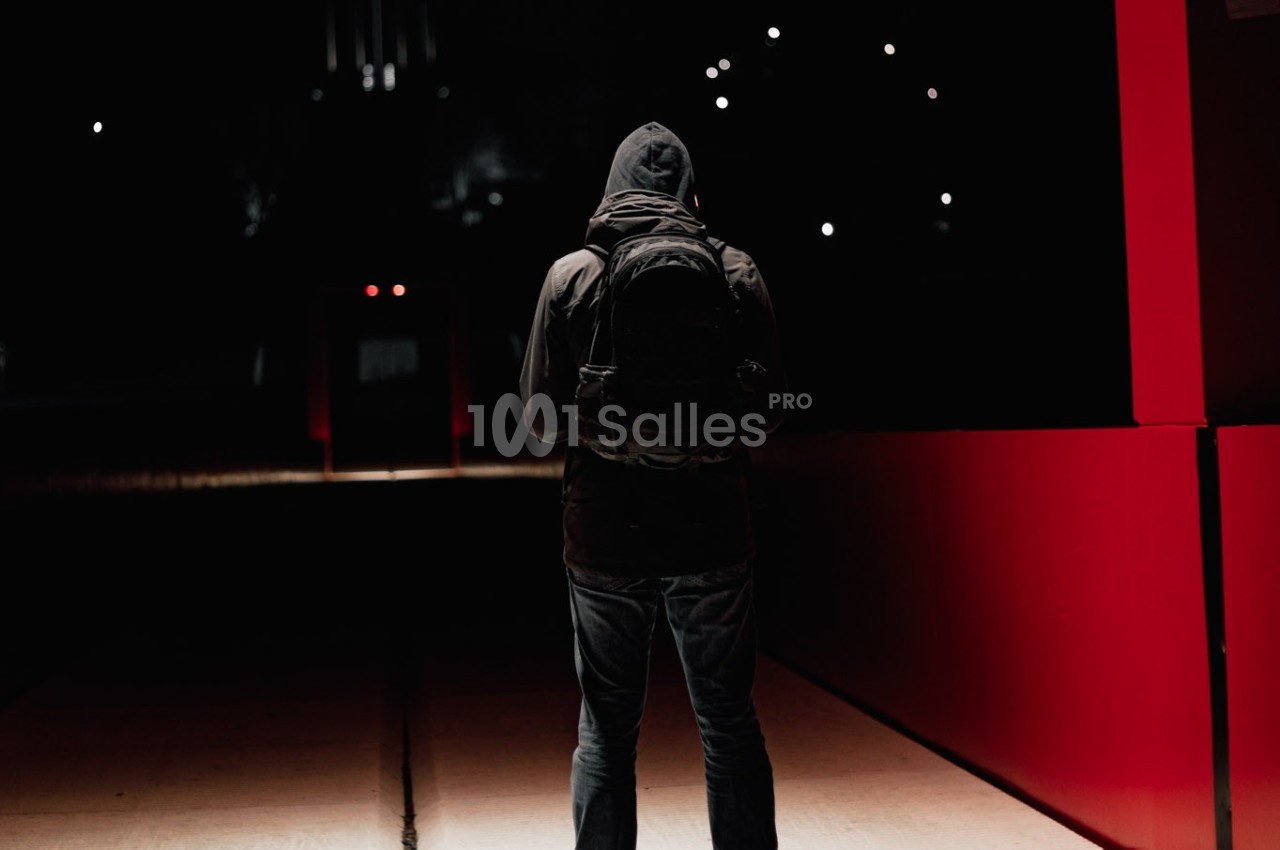 Un individu en manteau sombre et sac à dos, vu de dos, se tenant dans un espace faiblement éclairé avec des murs rouges.