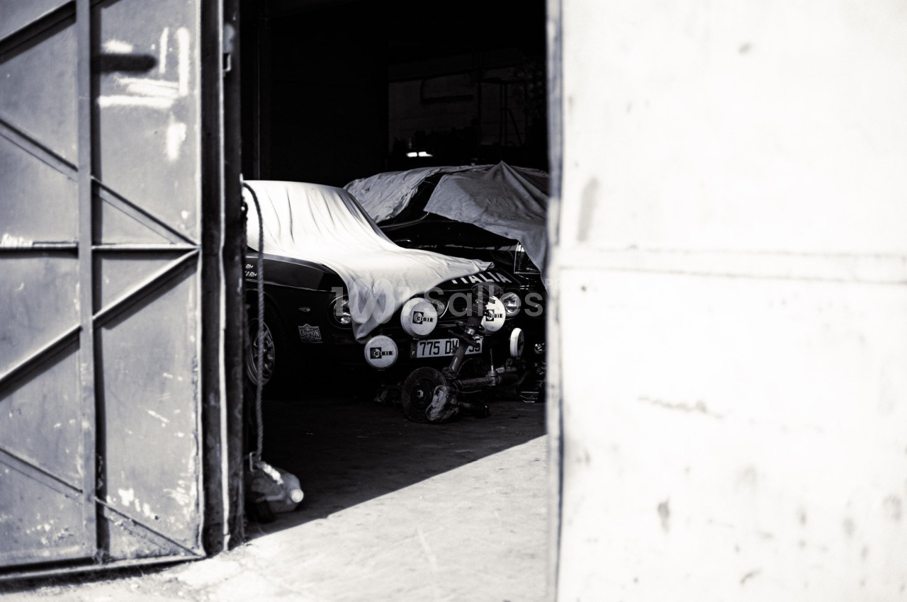 Voitures anciennes partiellement couvertes sous une bâche dans un garage sombre, vue à travers une porte entrouverte.