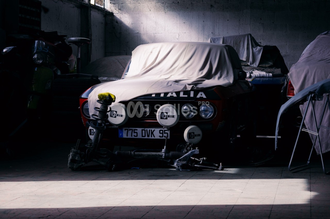 Voiture ancienne partiellement couverte dans un garage sombre, entourée d'équipements et d'autres véhicules.