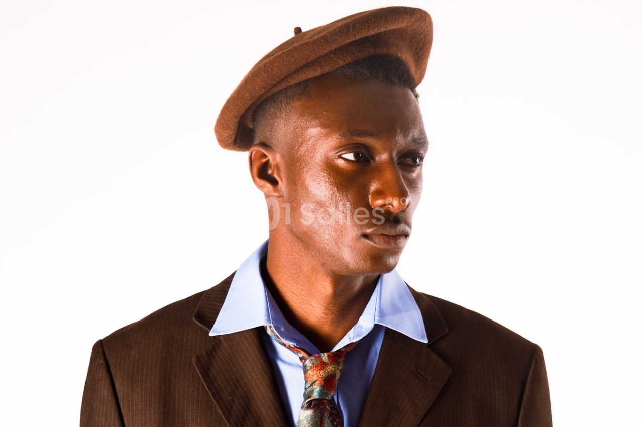 Homme portant un béret marron, une chemise bleue et une veste marron, avec une cravate légèrement dénouée.