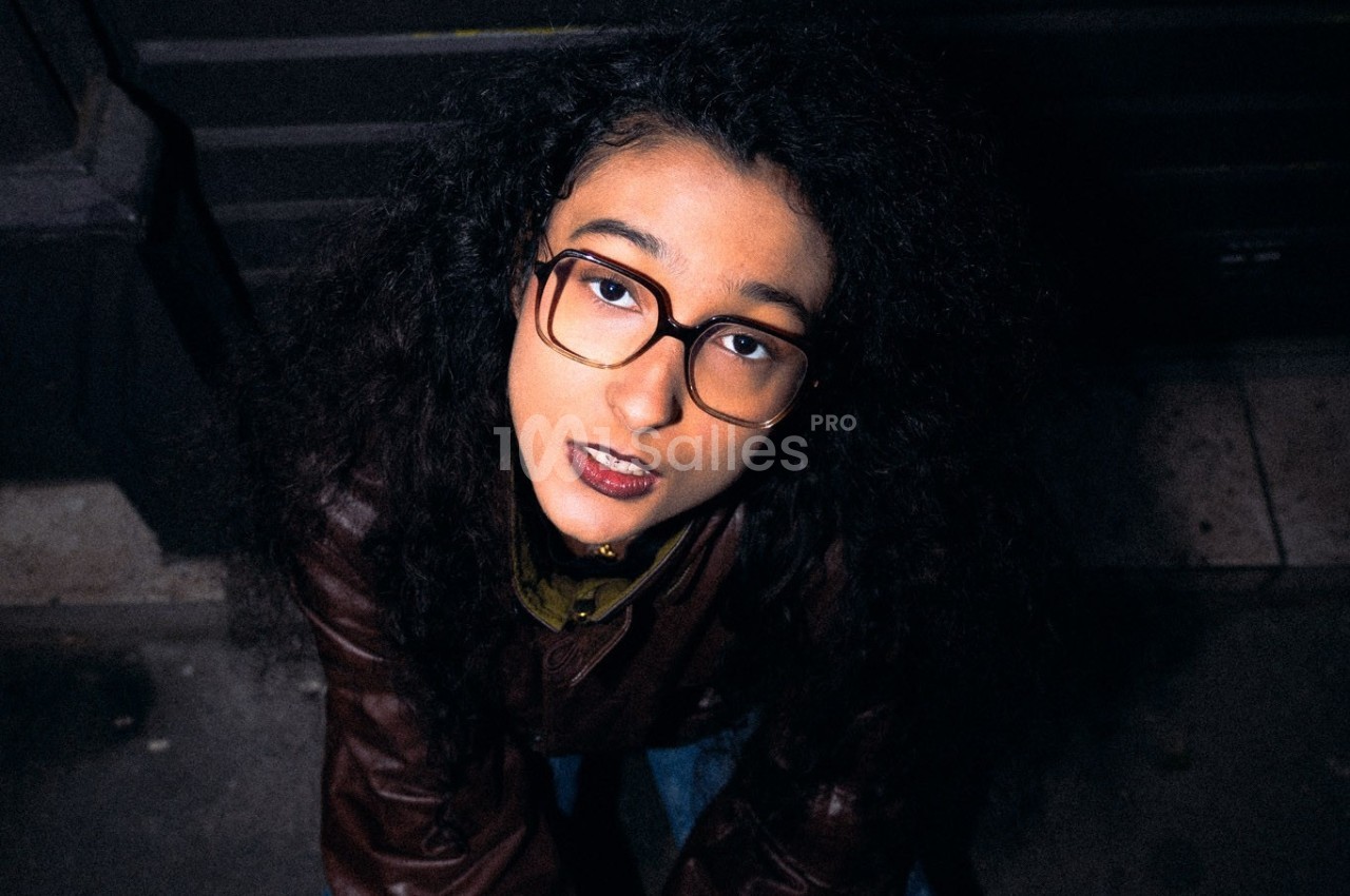 Femme aux cheveux bouclés et lunettes regardant vers le haut, vêtue d'une veste marron, en extérieur sombre.
