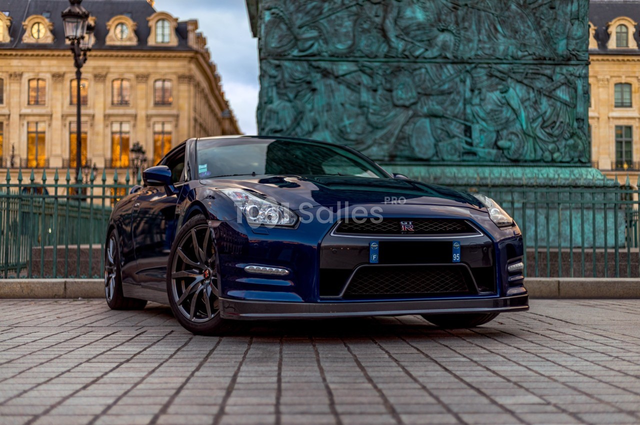 Voiture sportive bleue garée sur une place pavée devant des bâtiments historiques et une colonne monumentale.