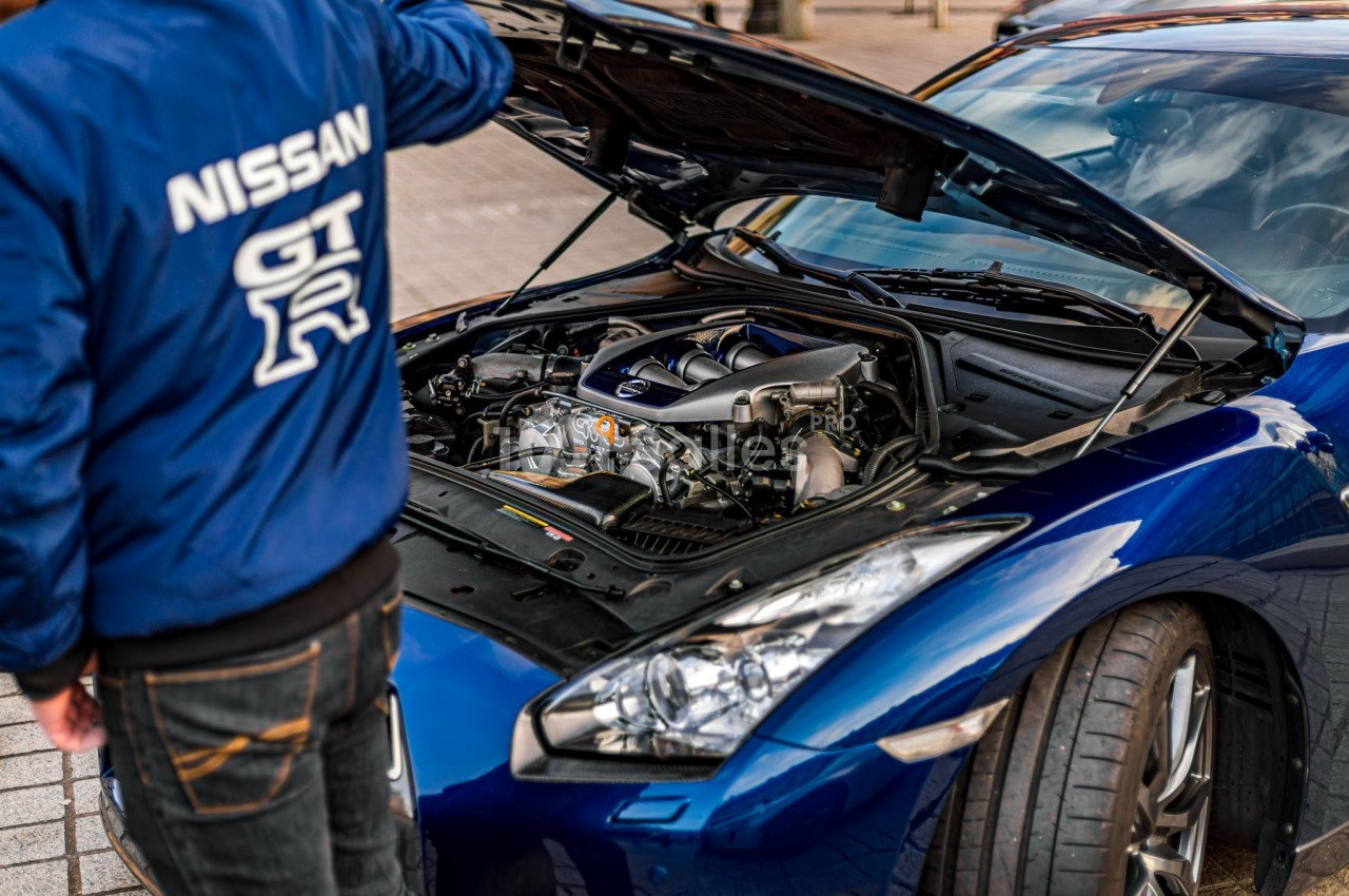 Un homme examine le moteur d'une voiture sportive bleue avec le capot ouvert, dans un environnement urbain.