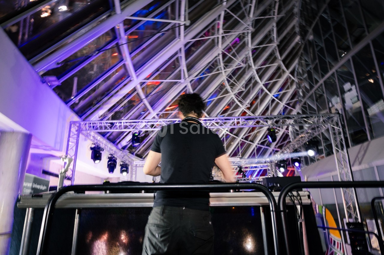 Un DJ de dos mixant sur une scène éclairée, dans un espace intérieur avec une structure en verre et métal.