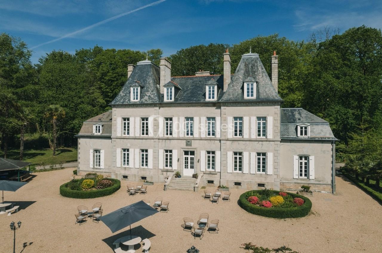 Façade d'un château en pierre entouré de jardins, avec des tables et parasols installés sur une cour gravillonnée.