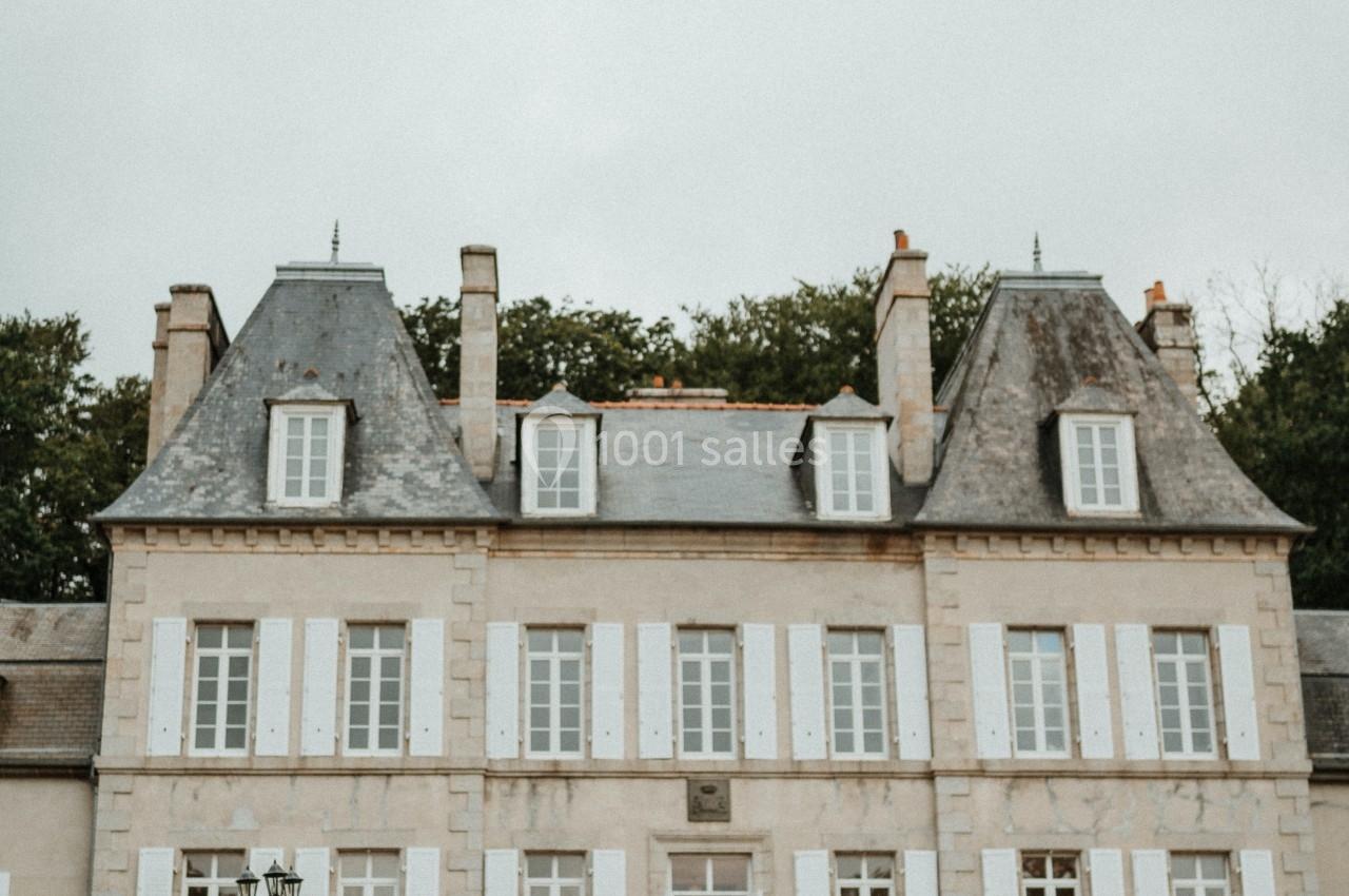 Un couple de mariés marche vers un château en pierre avec des volets blancs, entouré de quelques invités.