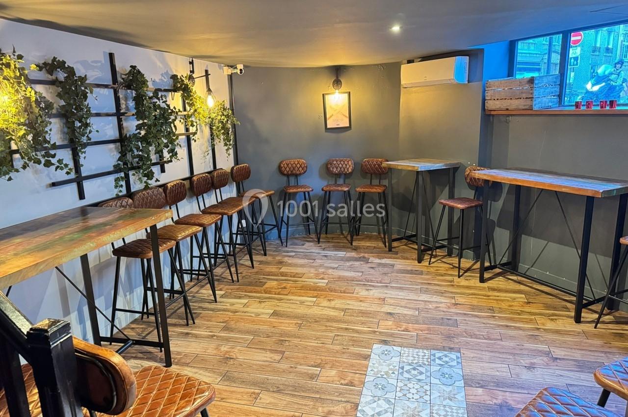 Salle lumineuse avec parquet, murs gris, tabourets en cuir, tables en bois et plantes suspendues en décoration.