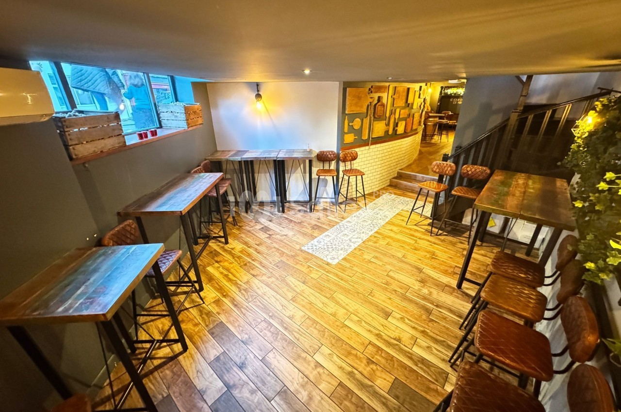 Salle lumineuse avec parquet en bois, tables hautes et tabourets, décorée de plantes et éclairée par des appliques murales.