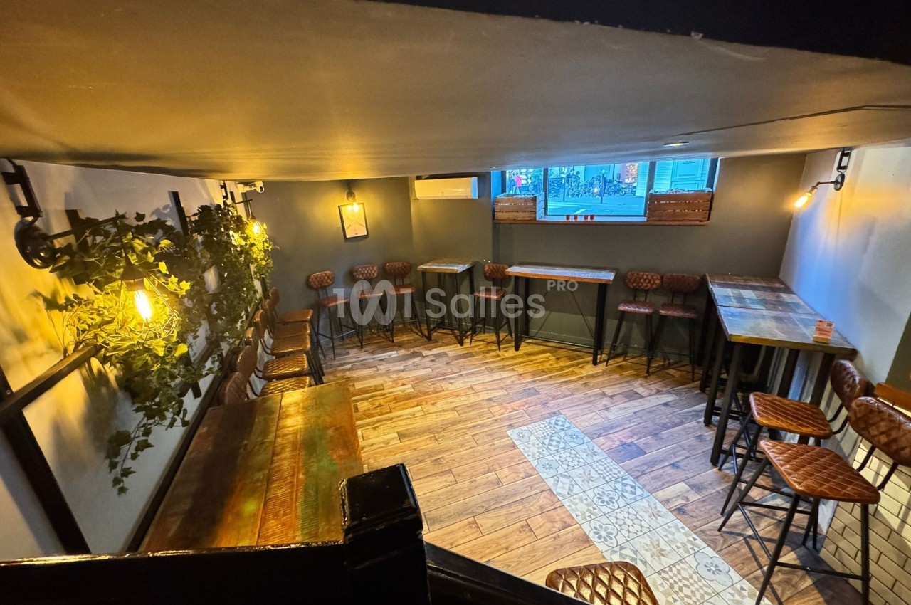 Salle lumineuse avec tables hautes, chaises en cuir, sol en bois et décorations murales végétales.