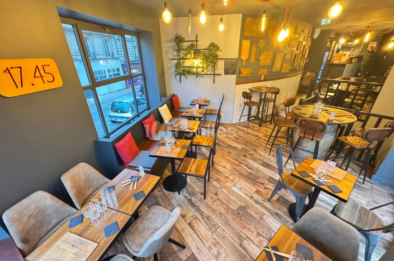 Salle de restaurant lumineuse avec tables en bois, chaises rembourrées, décoration murale et grandes fenêtres.