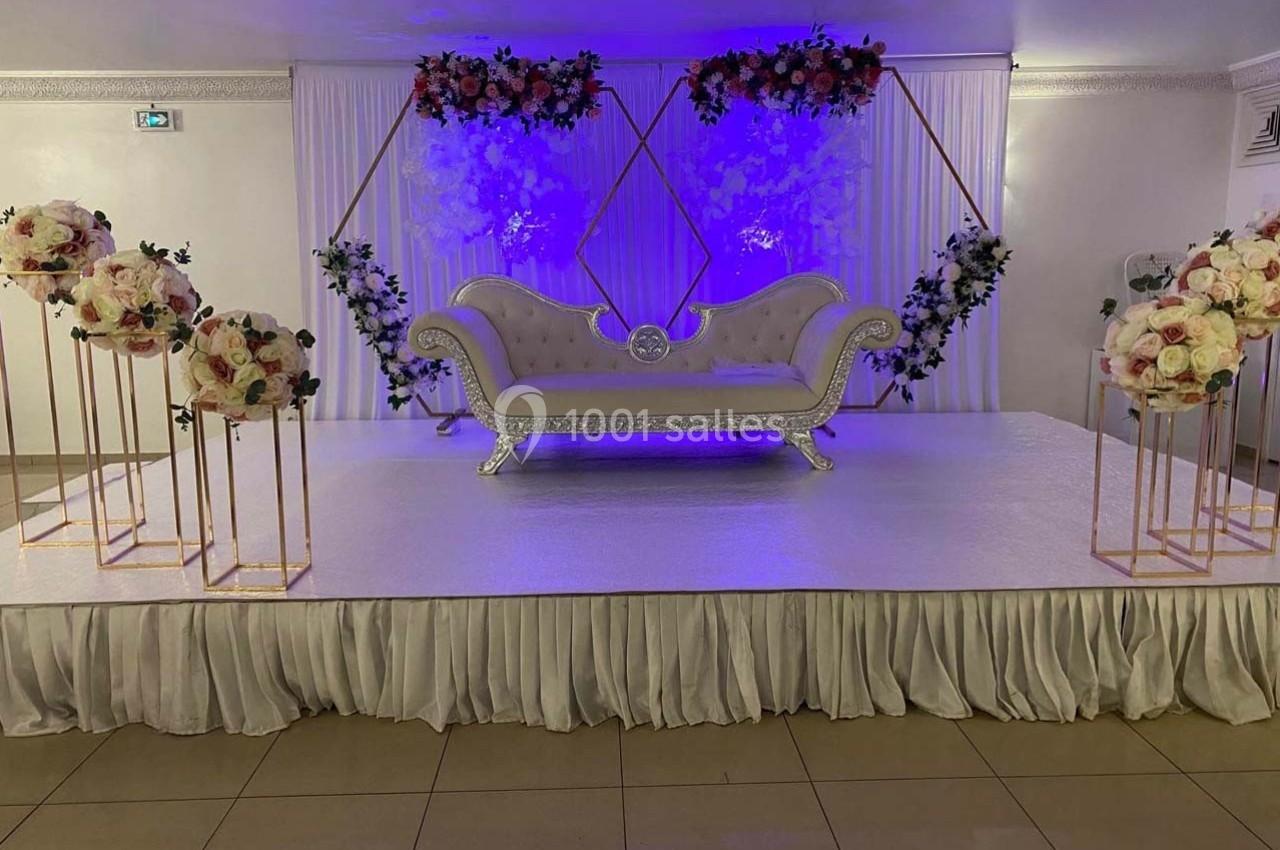 Canapé décoratif sur une scène ornée de fleurs et éclairée par une lumière violette dans une salle de réception.