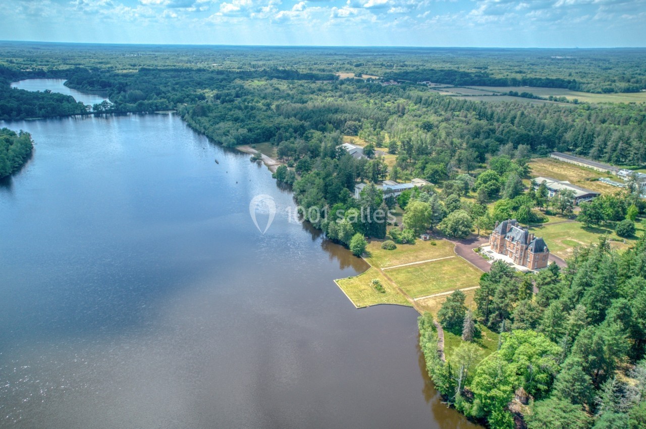 Vue aérienne d'un lac entouré de forêts et de prairies, avec un bâtiment en bordure et un ciel dégagé.