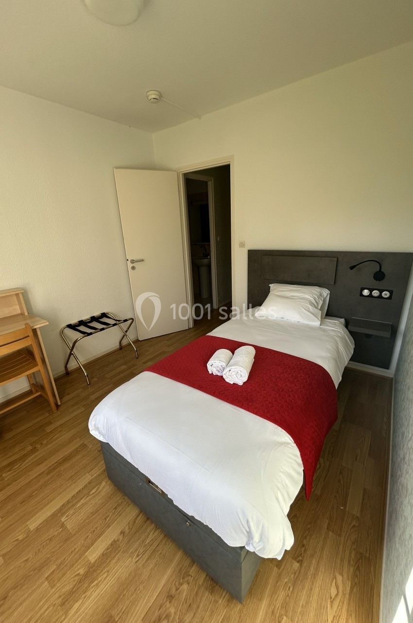 Chambre avec un lit simple, drap rouge, serviettes pliées, bureau en bois, sol en parquet et porte ouverte sur un couloir.