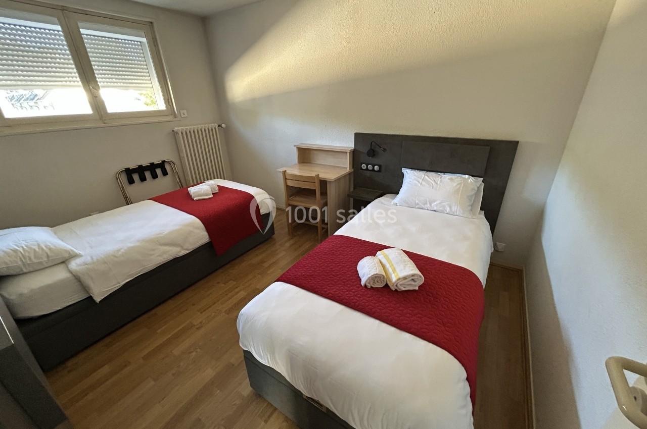Deux lits simples avec draps blancs et couvre-lits rouges dans une chambre lumineuse au sol en parquet.