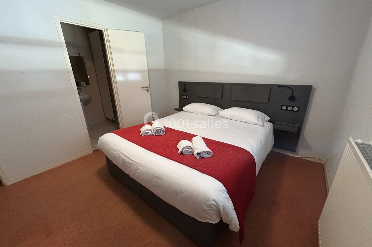 Chambre avec un lit double, draps blancs, couverture rouge, serviettes pliées et accès à une salle de bain adjacente.